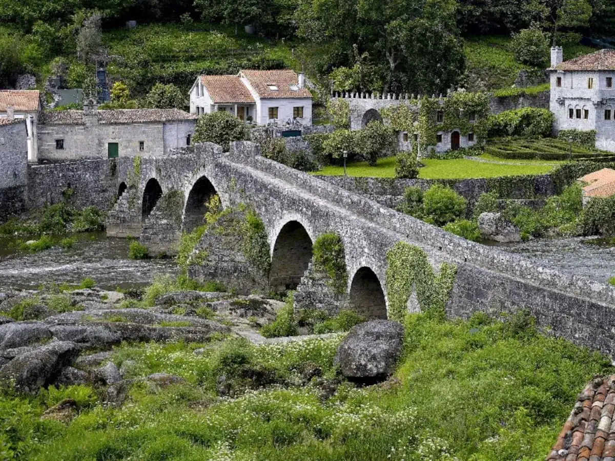 Un antiguo ponte carreira de piedra cruza un río, rodeado de casas rústicas y exuberante vegetación.