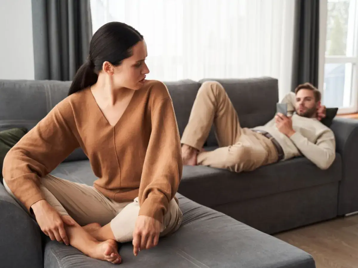 Une femme regarde son partenaire, qui est allongé sur le canapé, absorbé par son téléphone. La scène illustre la psychologie du refus de communiquer dans un couple.