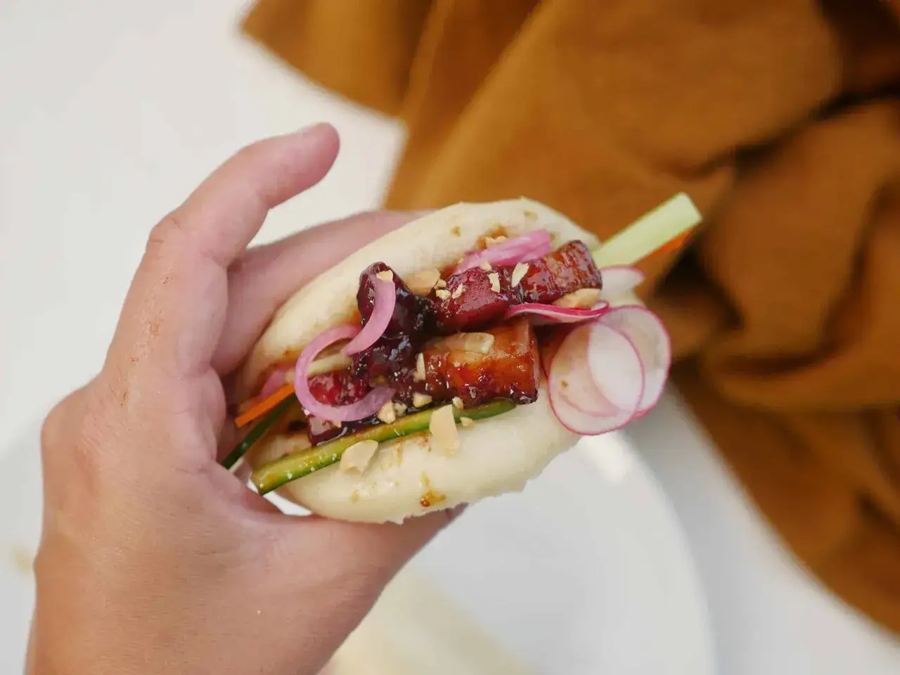 En hand håller en bao bun fylld med saftigt fläsk, krispig gurka, rödlök och jordnötter. Ett läckert bao buns recept!