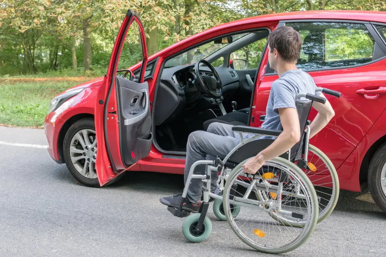 Un uomo in carrozzina si avvicina a un'auto rossa. La portiera è aperta, pronta per il trasferimento. Un disabile può avere due auto intestate per maggiore autonomia.