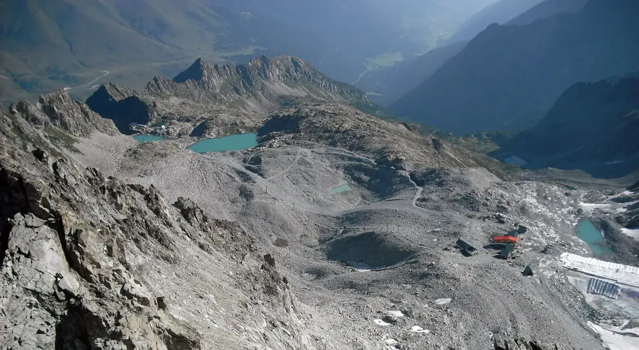 Un sentiero della pace serpeggia tra rocce e laghi alpini turchesi, con un rifugio rosso in lontananza.