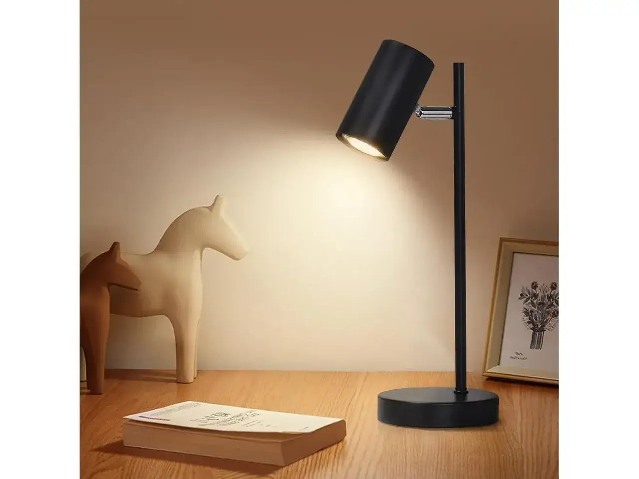 Una lampara mesilla negra ilumina un libro y dos figuras de caballos sobre una mesa de madera.