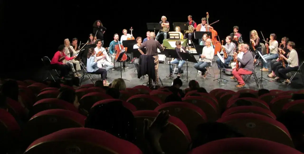 Un orchestre joue sur scène, dirigé par un chef. Les musiciens, dont certains jouent du violon, sont assis sur des chaises. L'ambiance rappelle une répétition pour la convention collective **idcc 1285**.