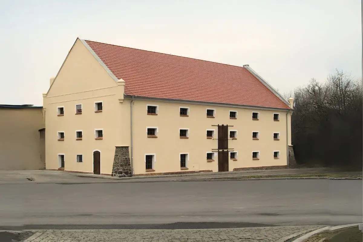 Jasny spichlerz w Jarocinie z czerwonym dachem i wieloma małymi oknami. Obok rosną drzewa.
