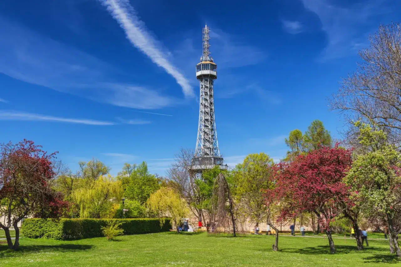 La tour Eiffel de Prague, une structure m&eacute;tallique imposante, se dresse fi&egrave;rement au-dessus d'un parc verdoyant et fleuri sous un ciel bleu &eacute;clatant.