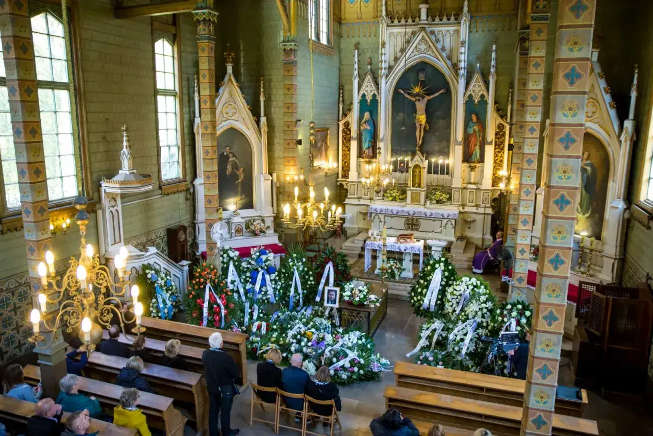 Wnętrze kościoła podczas ceremonii pogrzebowej urny. Wiele wieńców pogrzebowych otacza miejsce pochówku.