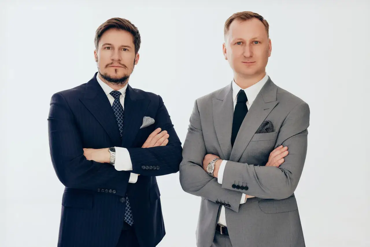 Dwóch mężczyzn w garniturach, stojących z rękami skrzyżowanymi na piersi.