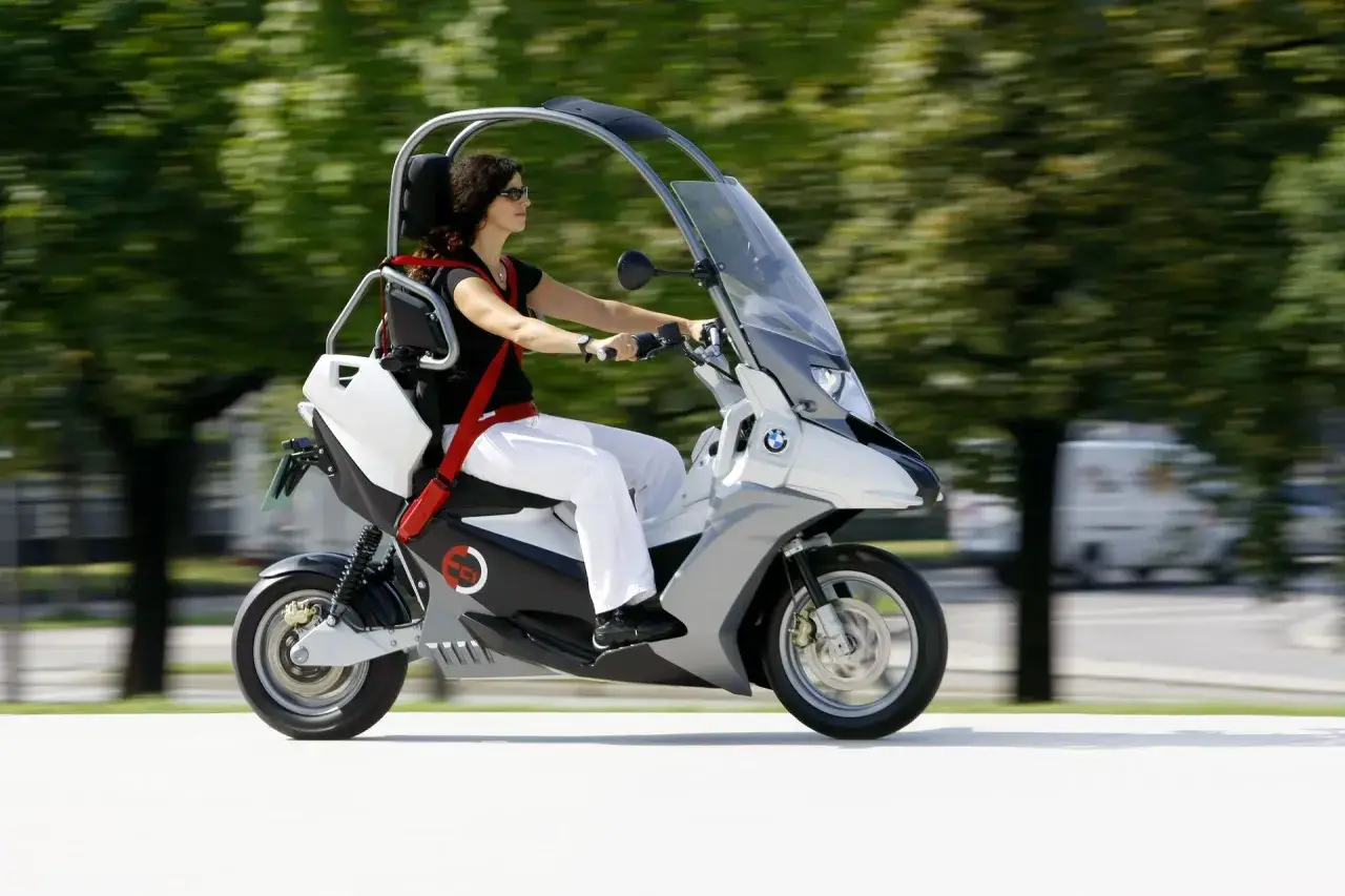 Kobieta w białych spodniach i czarnej bluzce jedzie na futurystycznym skuterze BMW C 400 X z metalową ramą i dachem.