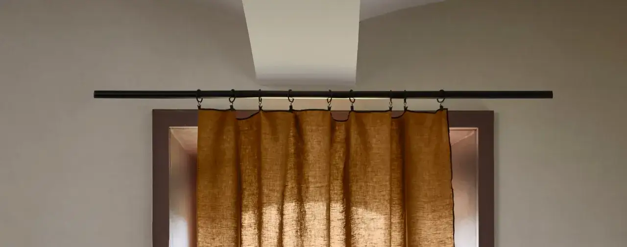 Une tringle à rideaux noire est fixée au mur au-dessus d'une fenêtre. Des anneaux noirs sont accrochés à la tringle, supportant un rideau en lin brun.