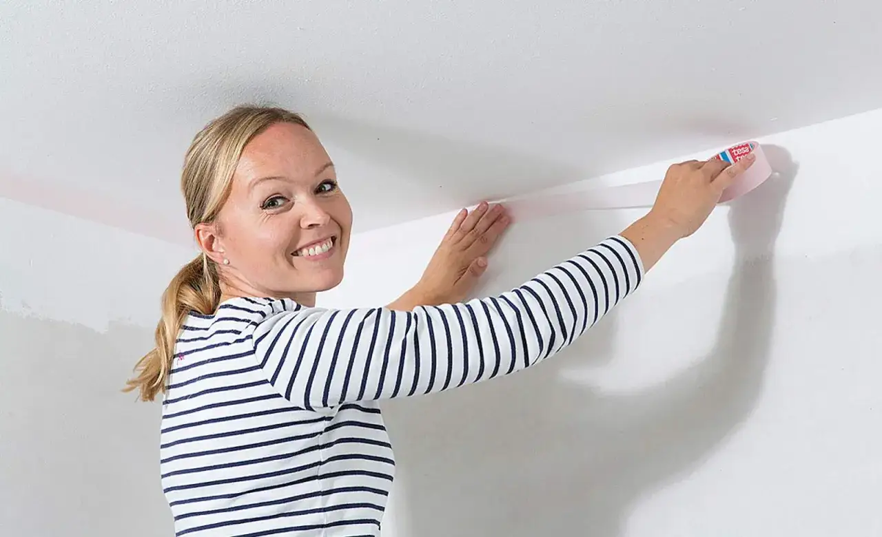 Frau klebt Klebeband an die Decke, um eine gerade Kante für das zweifarbige Wand streichen zu schaffen.