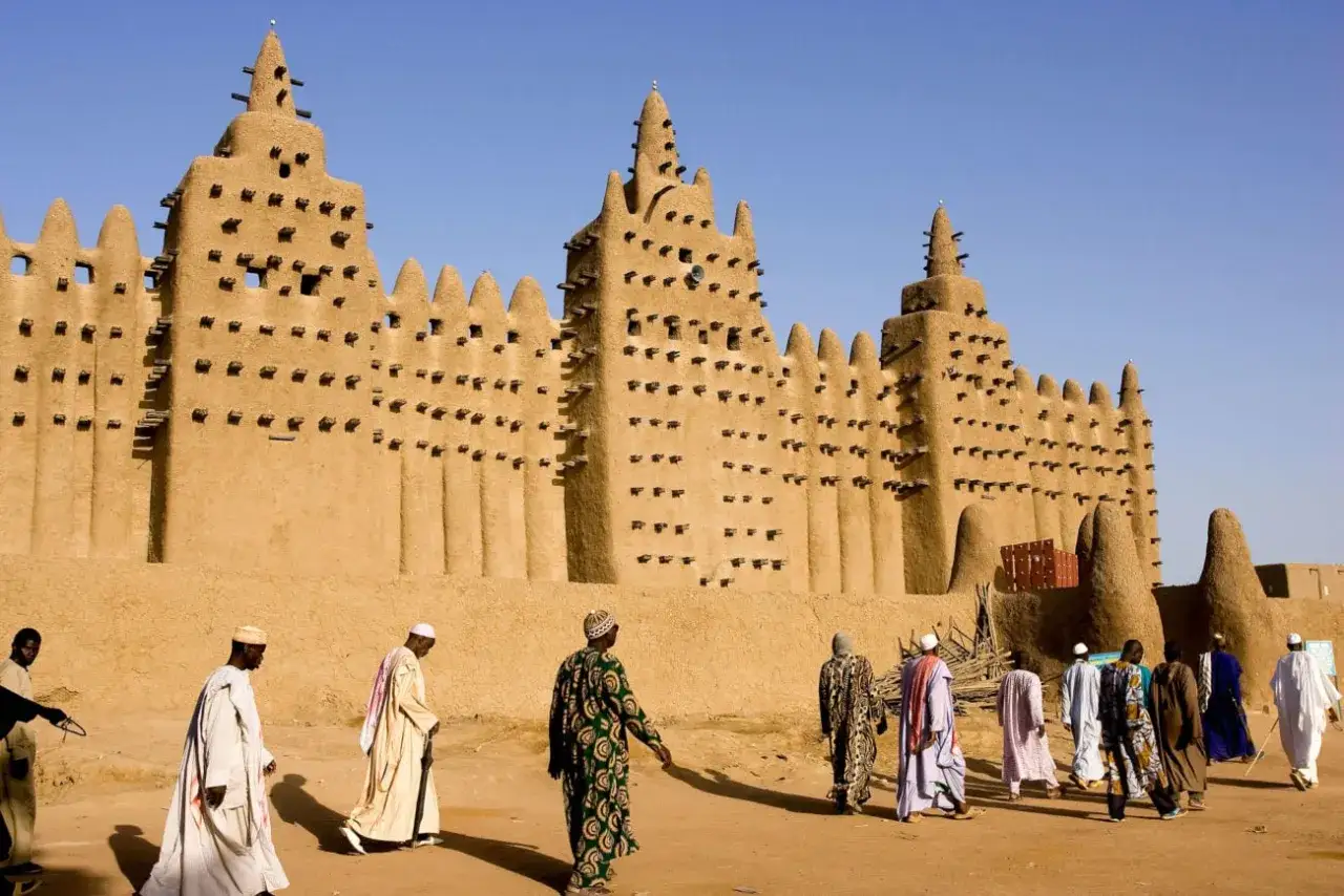 La grande mosqu&eacute;e de Djenn&eacute;, chef-d'&oelig;uvre d'architecture en terre, se dresse sous un ciel bleu. Des hommes en tenues traditionnelles s'approchent, ajoutant vie &agrave; ce site historique.