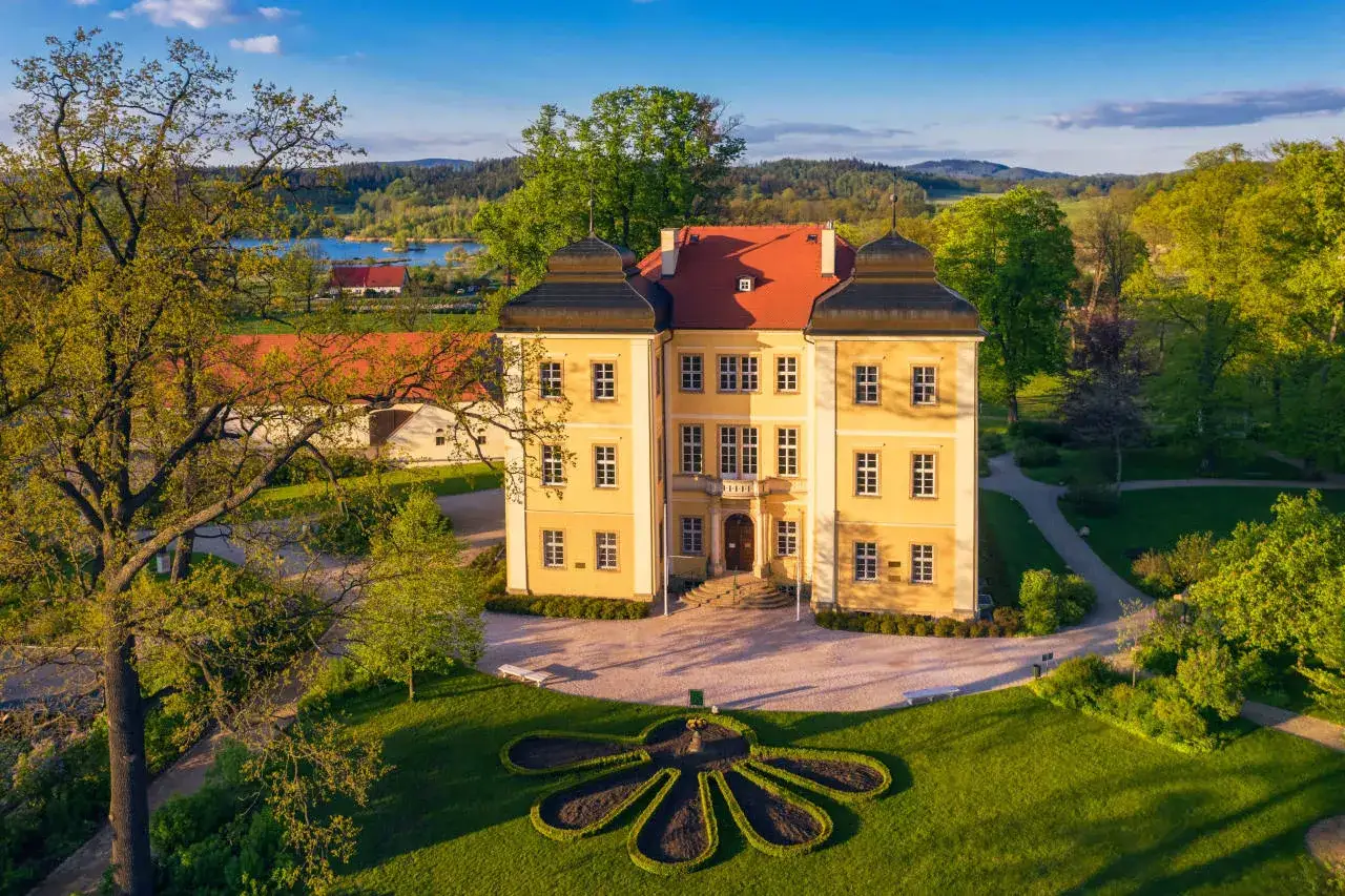 Pałac Łomnica zaprasza na zwiedzanie. Majestatyczna budowla otoczona zielenią, z jeziorem w tle.