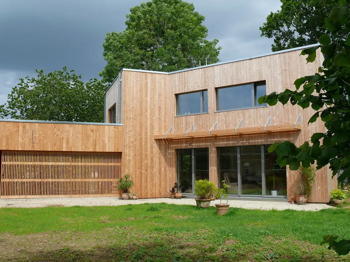 Une maison passive moderne en bois, avec de grandes baies vitr&eacute;es et une porte de garage ajour&eacute;e, entour&eacute;e de verdure.