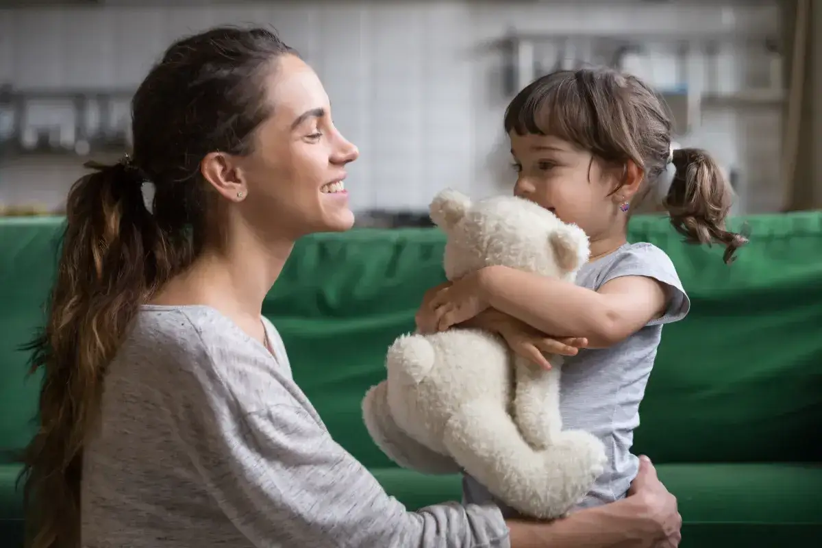Uśmiechnięta mama patrzy na córkę tulącą misia. Czas spędzony razem, jak ten, zasługuje na zasiłek opiekuńczy.
