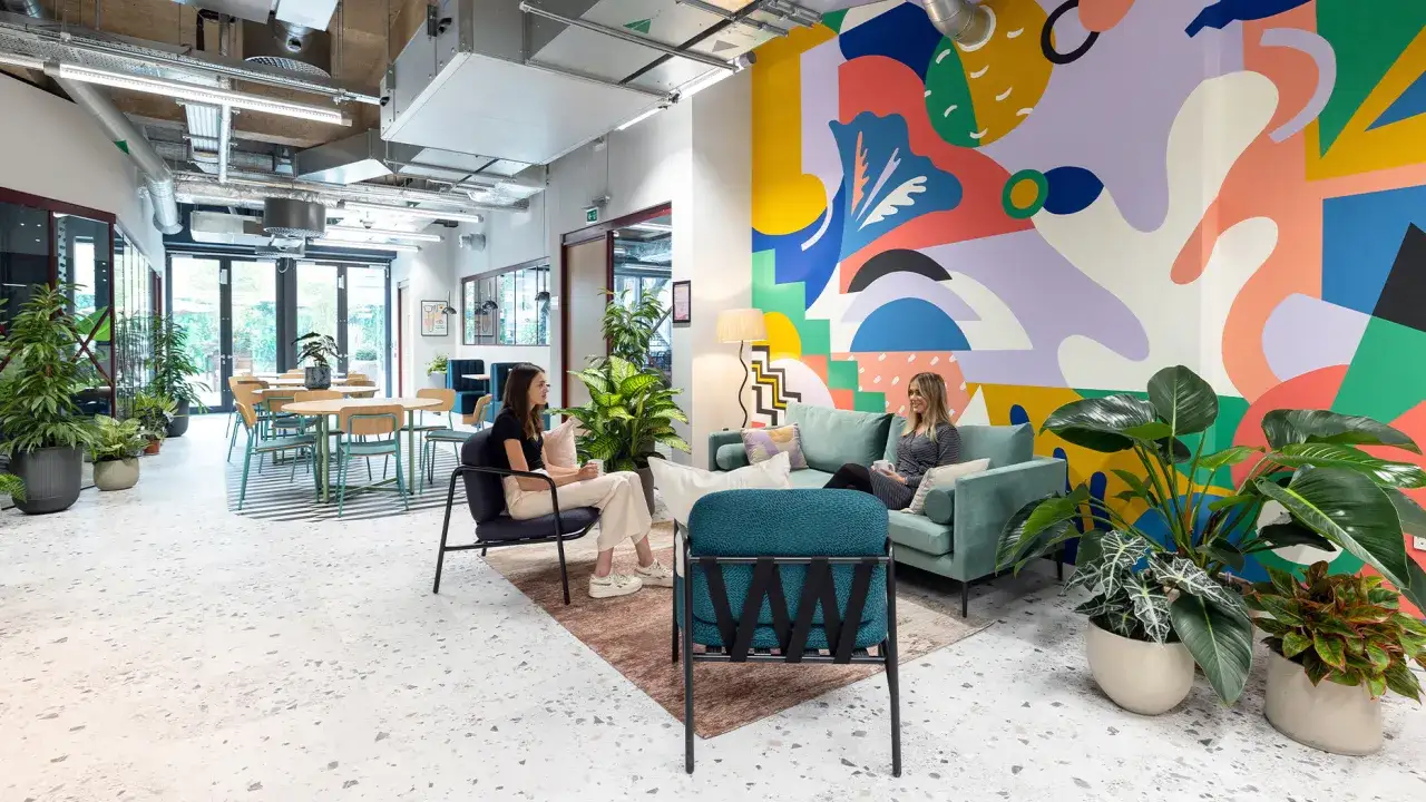 Aménagement espace coworking coloré avec mur graphique, plantes vertes et deux femmes discutant sur un canapé.