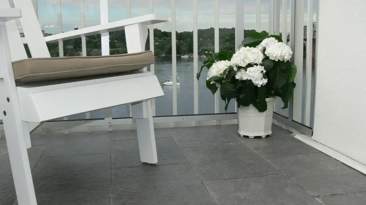 En vit stol och en hortensia på en balkong med utsikt över vatten. Kakelfix utomhus ger en stabil grund för utemöbler.