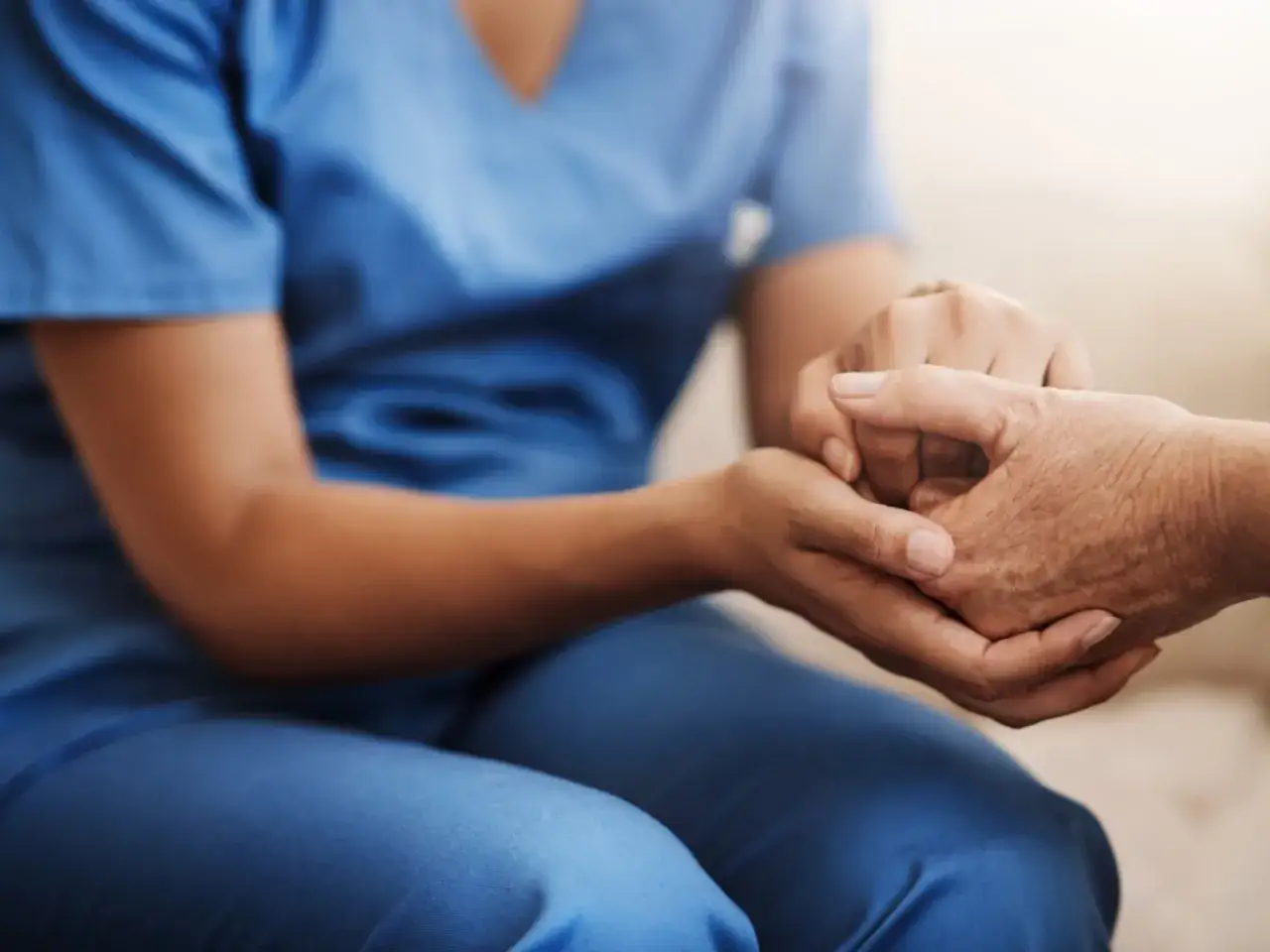 Opiekun medyczny w niebieskim uniformie trzyma dłoń starszej osoby. To pokazuje, gdzie może pracować opiekun medyczny: w domach, szpitalach, hospicjach.