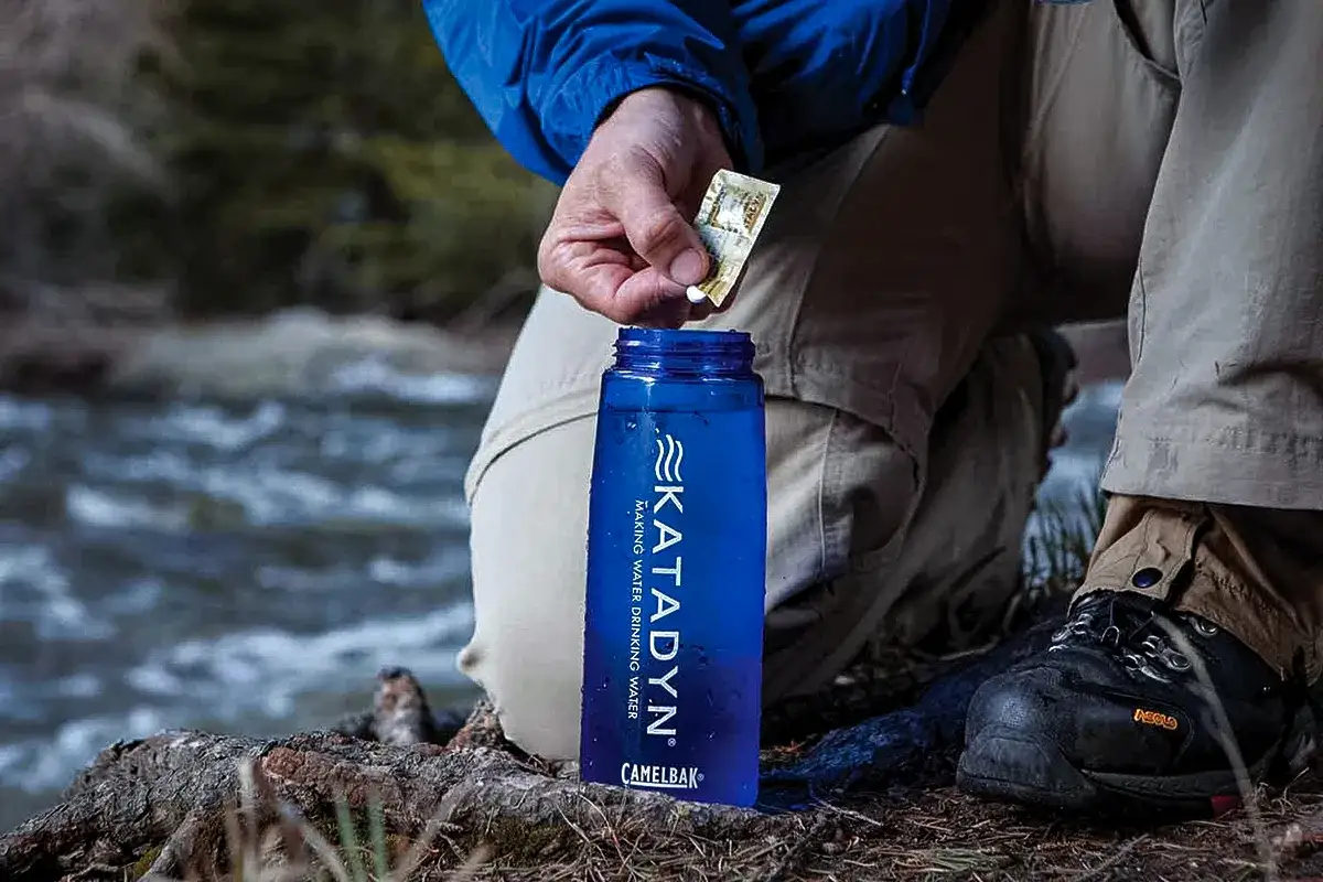 Persona a&ntilde;ade una pastilla Katadyn Micropur Forte a una botella de agua azul junto a un r&iacute;o.
