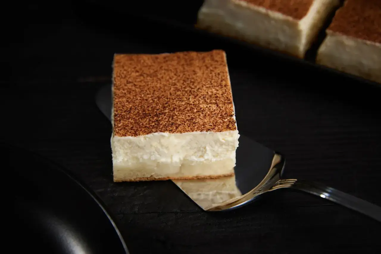 Ein Stück cremiger Rahmkuchen vom Blech, bestäubt mit Kakaopulver, wird auf einem Tortenheber präsentiert.
