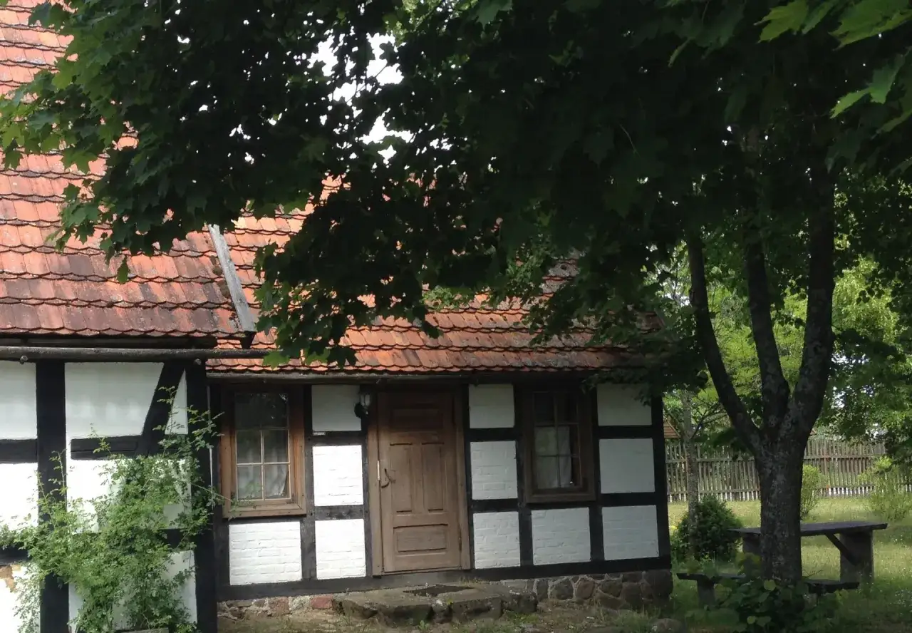 Urokliwa chata kaszubska z drewnianymi belkami i czerwoną dach&oacute;wką, otoczona zielenią.