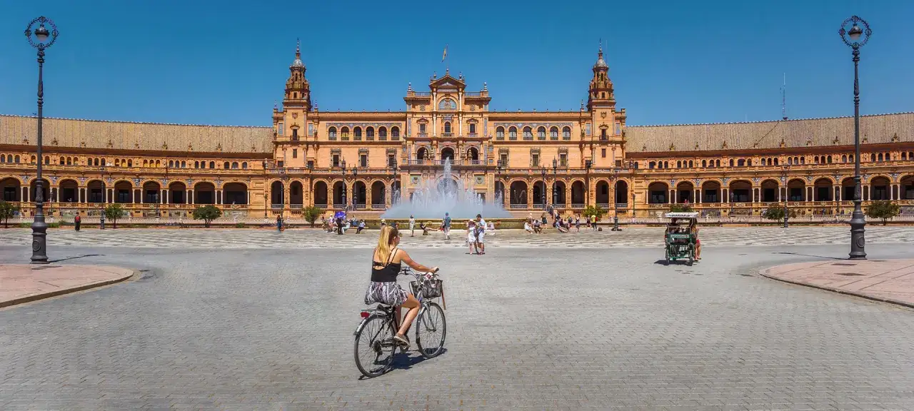 En kvinna cyklar på Plaza de España i Sevilla, Spanien, med en stor fontän och en imponerande byggnad i bakgrunden.