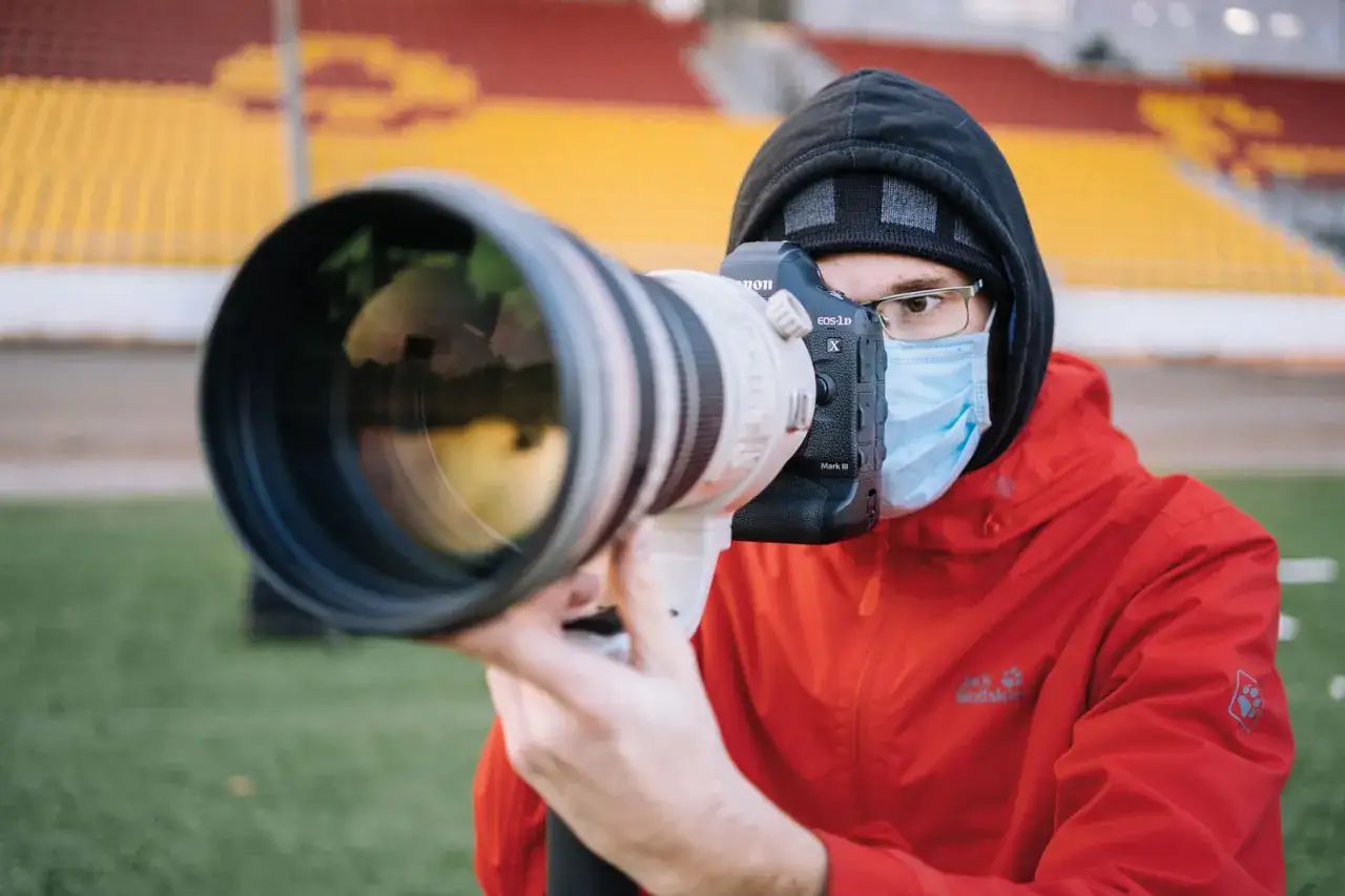 Fotograf sportowy w masce i czerwonej kurtce z profesjonalnym aparatem Canon EOS-1D X Mark III na tle trybun stadionu.