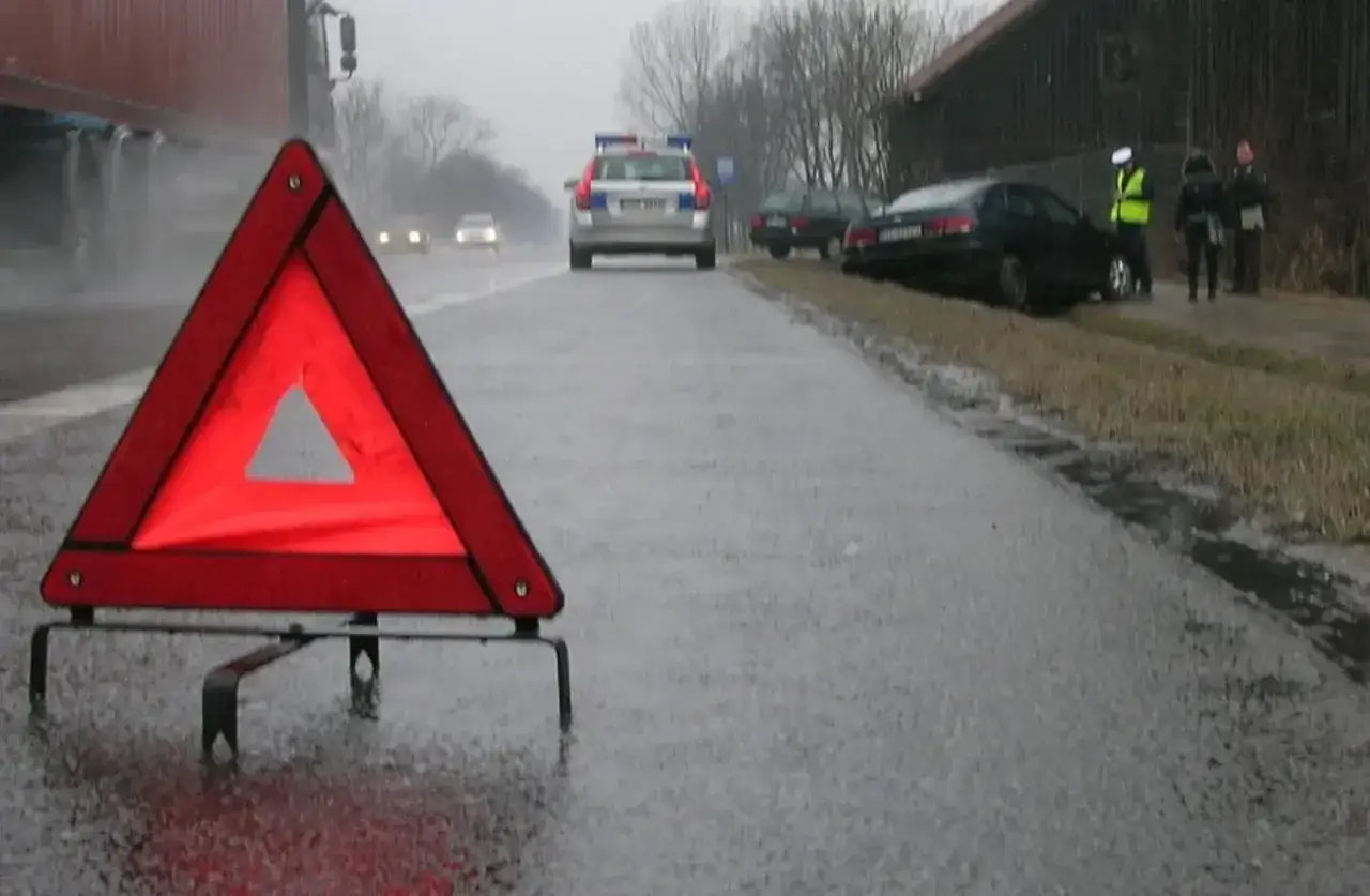 Czerwony trójkąt ostrzegawczy na mokrej drodze, obok radiowóz i osoby. W tle widać ciężarówkę i inne pojazdy. Światła awaryjne migają, ostrzegając przed niebezpieczeństwem.