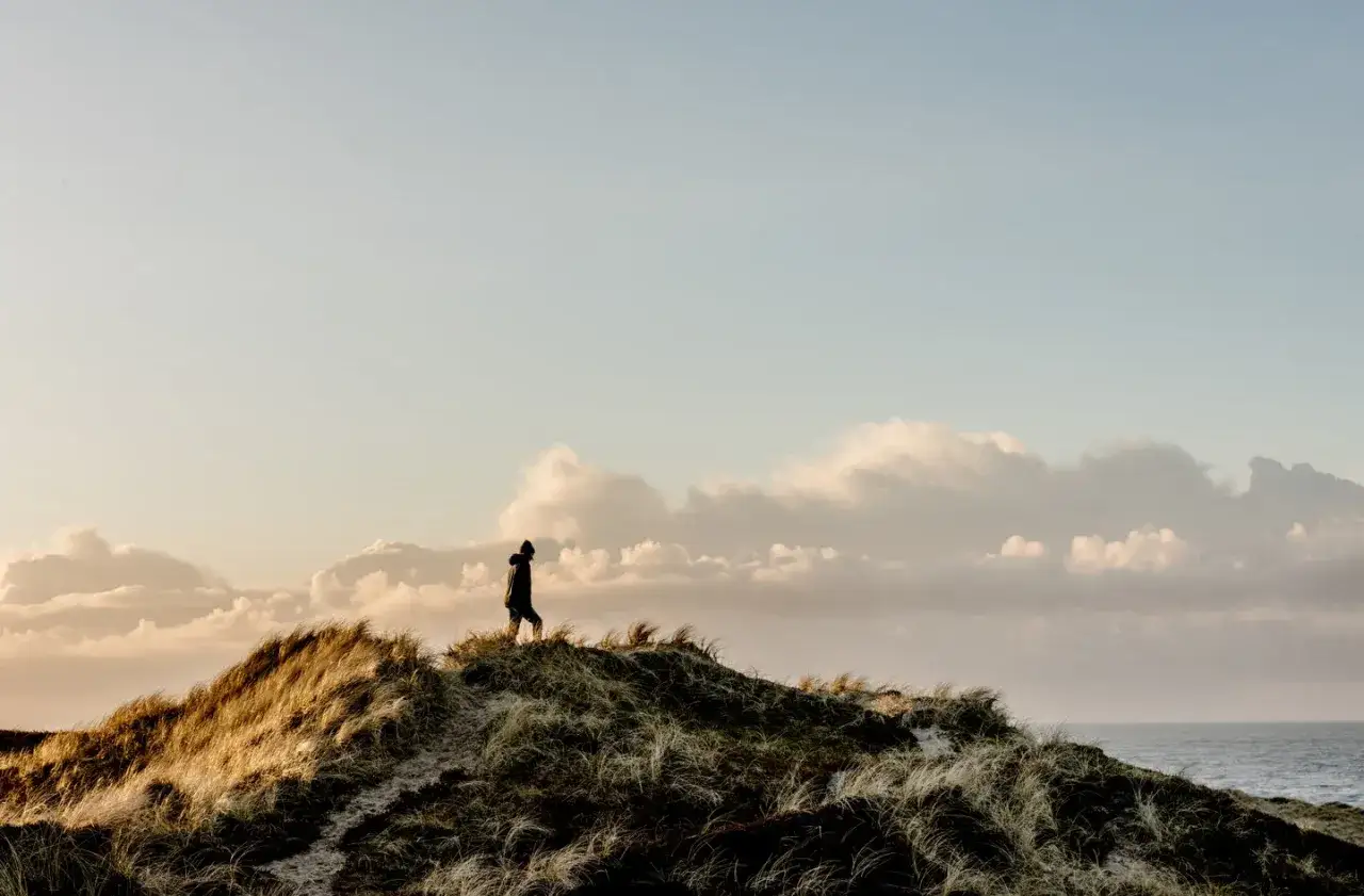En person g&aring;r p&aring; en sanddyn vid kusten i Thy, Danmark. Solen g&aring;r ner och himlen &auml;r fylld av moln.