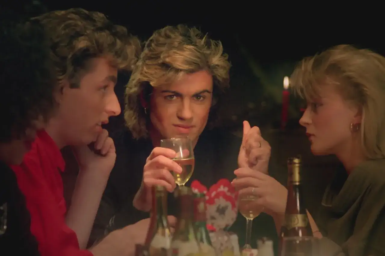 George Michael, który śpiewa Last Christmas, siedzi przy stole z przyjaciółmi, pijąc wino.