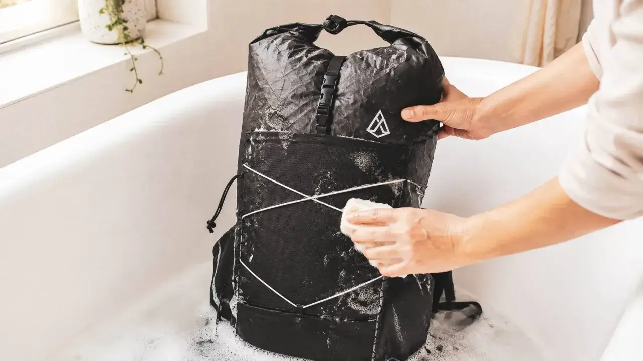Person reinigt einen schwarzen Rucksack in einer Badewanne mit Schaum. Der Rucksack wird mit einem Schwamm abgerieben, um ihn zu waschen.