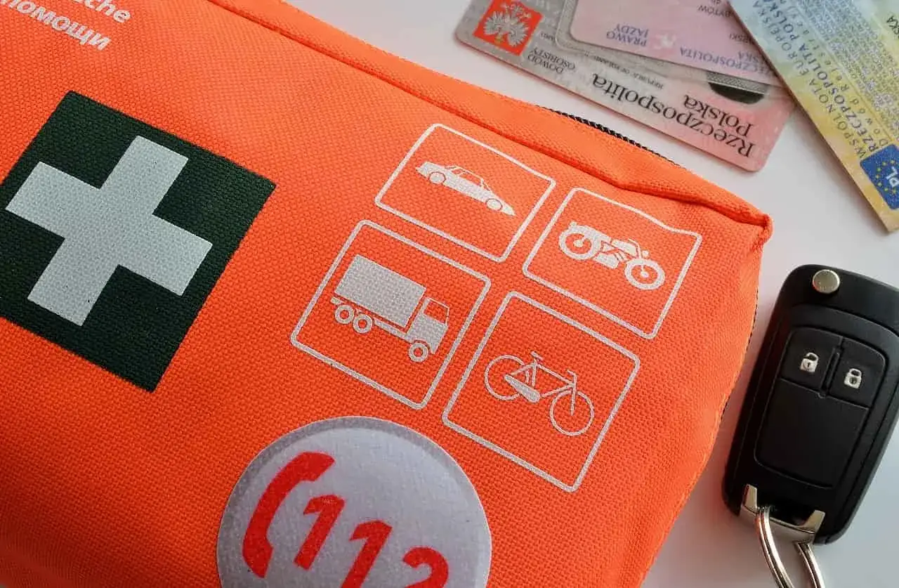 Pomarańczowa apteczka samochodowa z symbolem krzyża i ikonami pojazdów. Czy apteczka jest obowiązkowa w samochodzie? Kluczyki i dokumenty obok.
