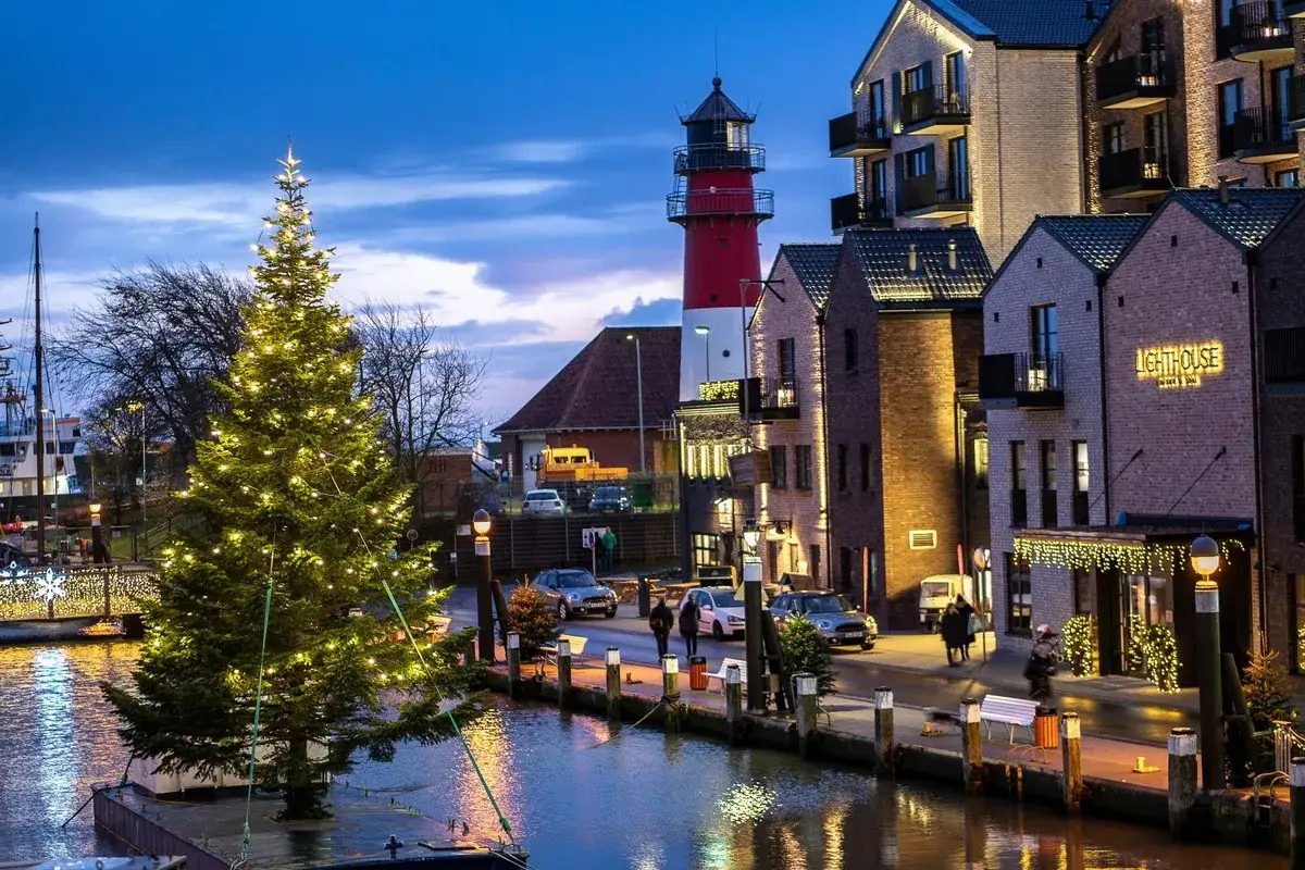 Ein festlich beleuchteter Weihnachtsbaum schwimmt im Wasser, während im Hintergrund ein Leuchtturm und weihnachtlich geschmückte Häuser die Atmosphäre eines Weihnachtsmarktes in Schleswig-Holstein zaubern.