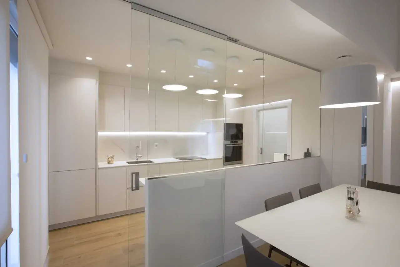 Cocina moderna con puertas de cristal correderas para separar cocina de sal&oacute;n. Muebles blancos, encimera de m&aacute;rmol y suelo de madera.
