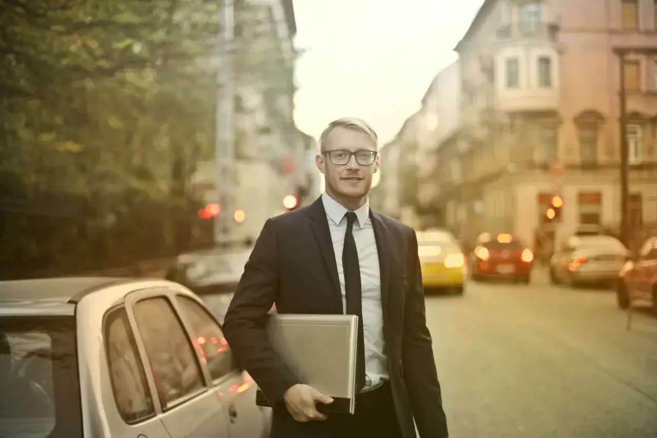 Uśmiechnięty przedstawiciel handlowy w garniturze, z laptopem w ręku, idzie ulicą. Jego zakres obowiązk&oacute;w obejmuje spotkania z klientami i prezentacje.