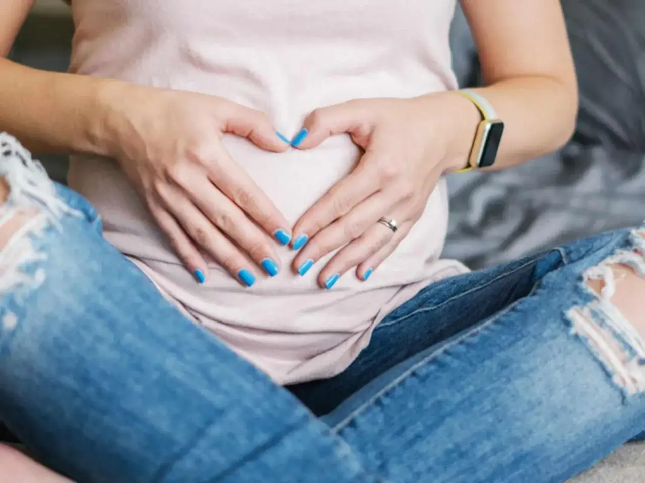 Schwangere Frau mit blauem Nagellack formt Herz auf Bauch. Ein Zeichen für die Vorfreude auf das Baby, auch mit Nagellack in der Schwangerschaft.