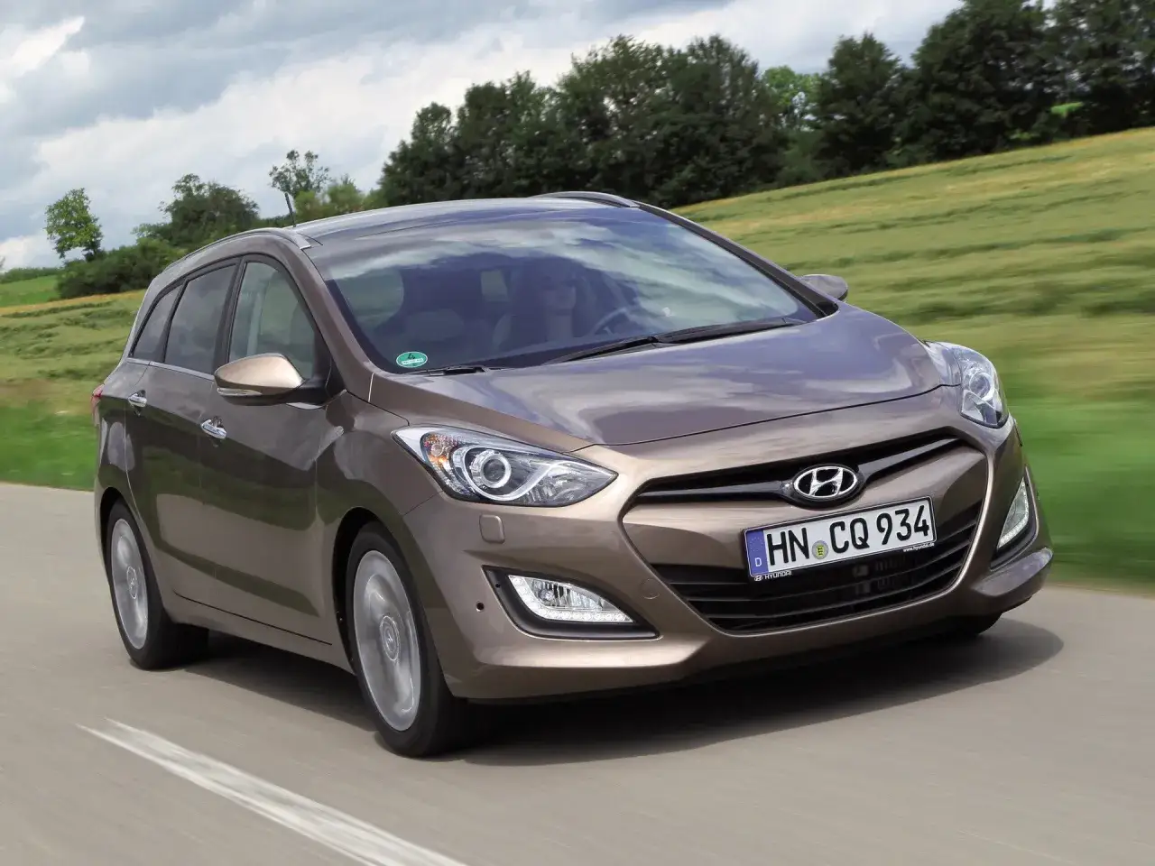 Brązowy Hyundai i30 1.6 benzyna w wersji kombi jedzie po drodze wśród zielonych pól.