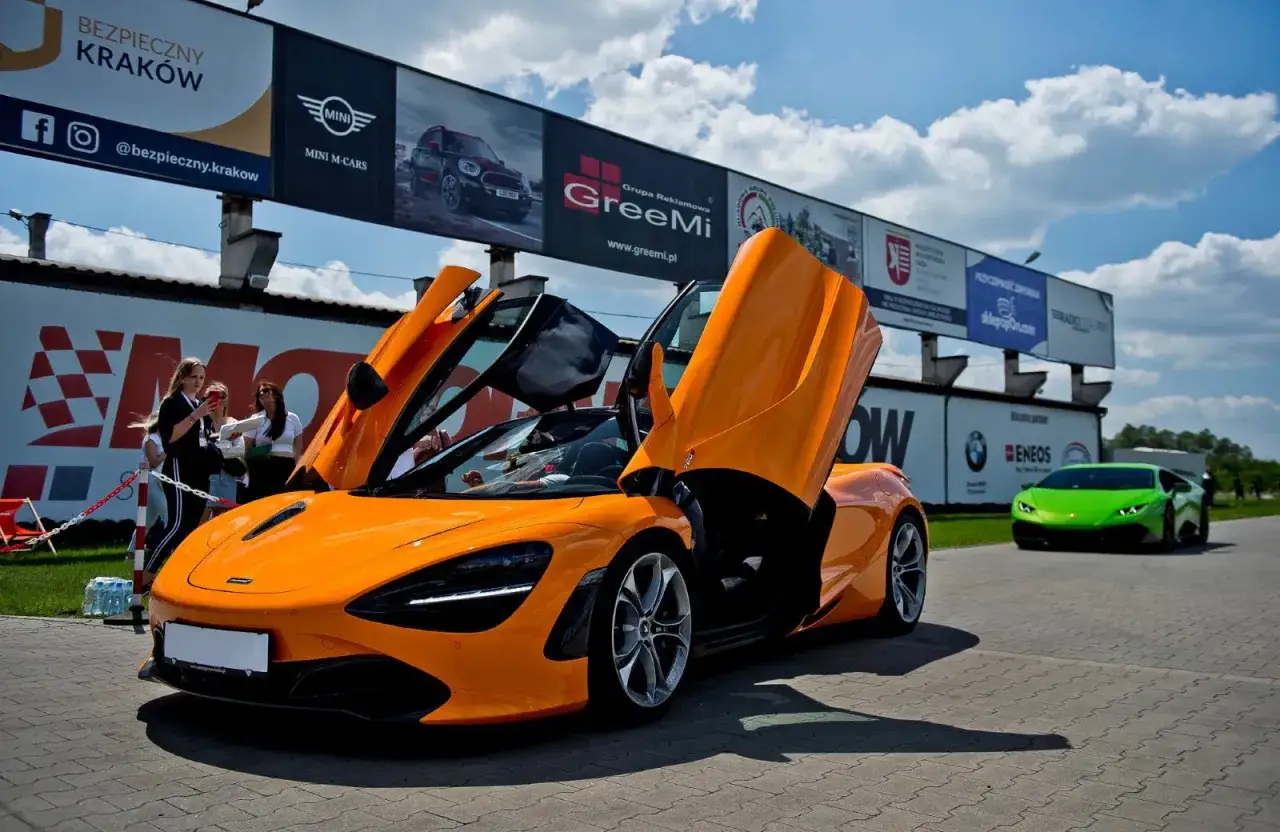 Pomarańczowy McLaren 720S z otwartymi drzwiami na torze wyścigowym, obok zielony Lamborghini.