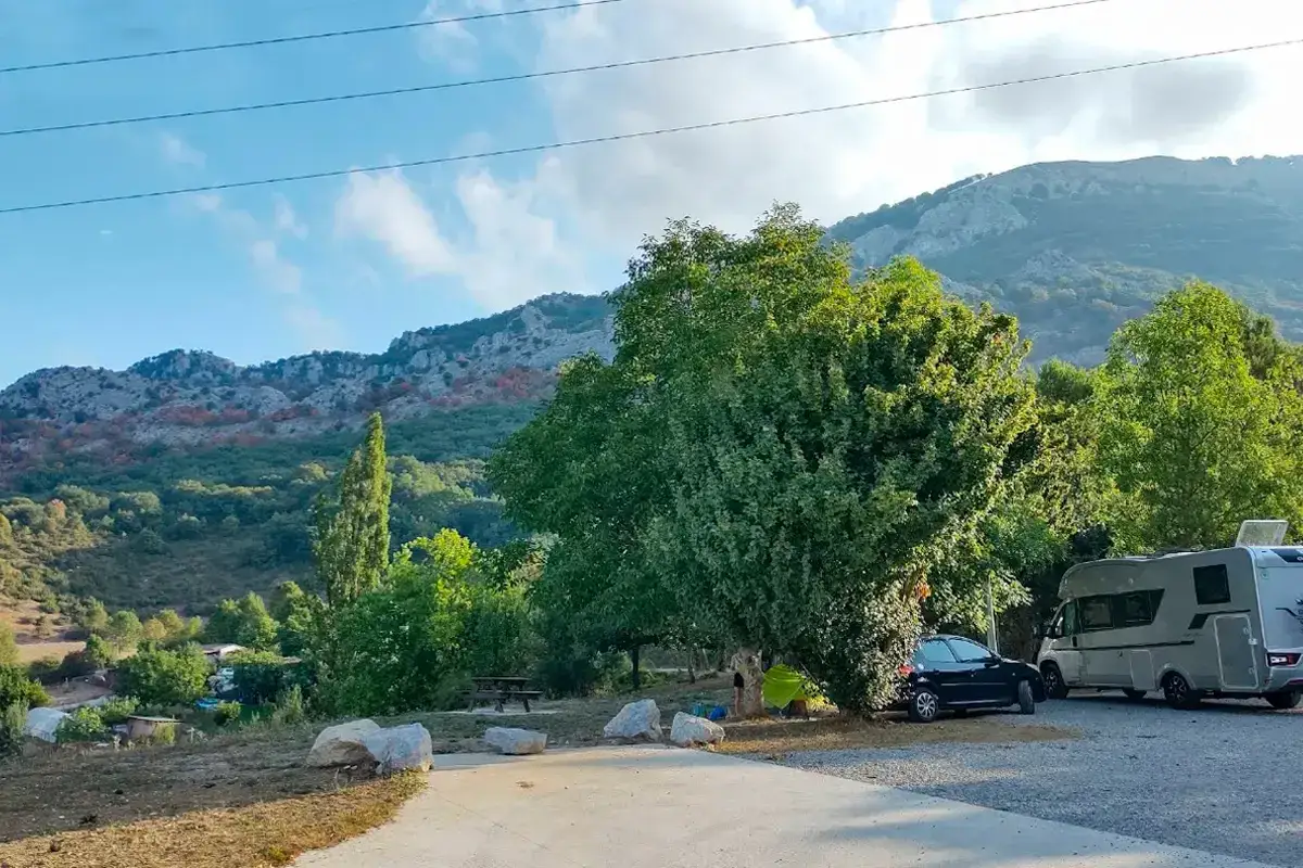 Un área camper en Pamplona con autocaravana y coche aparcados bajo árboles frondosos, con montañas de fondo.