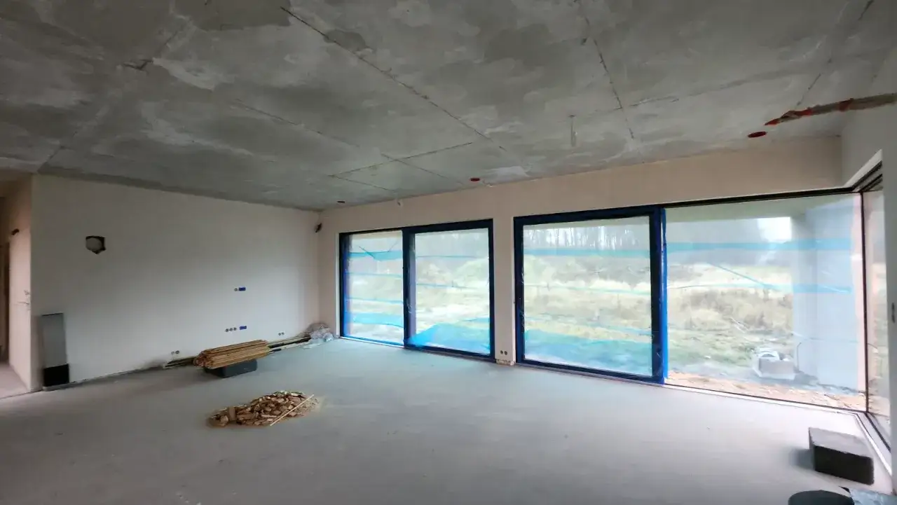 Wnętrze domu w stanie deweloperskim. Betonowy sufit, nieotynkowane ściany, wielkie okna z widokiem na las.