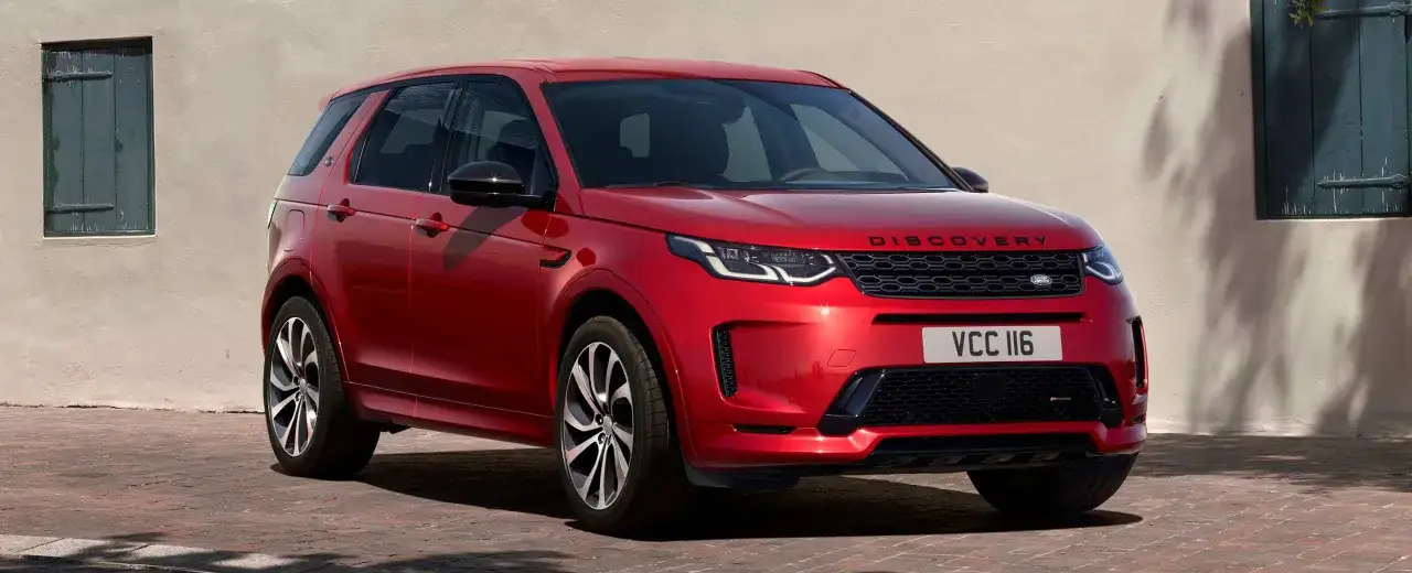 Roter Land Rover Discovery Sport, ein **land rover approved** Fahrzeug, steht vor einer hellen Wand mit gr&uuml;nen Fensterl&auml;den.