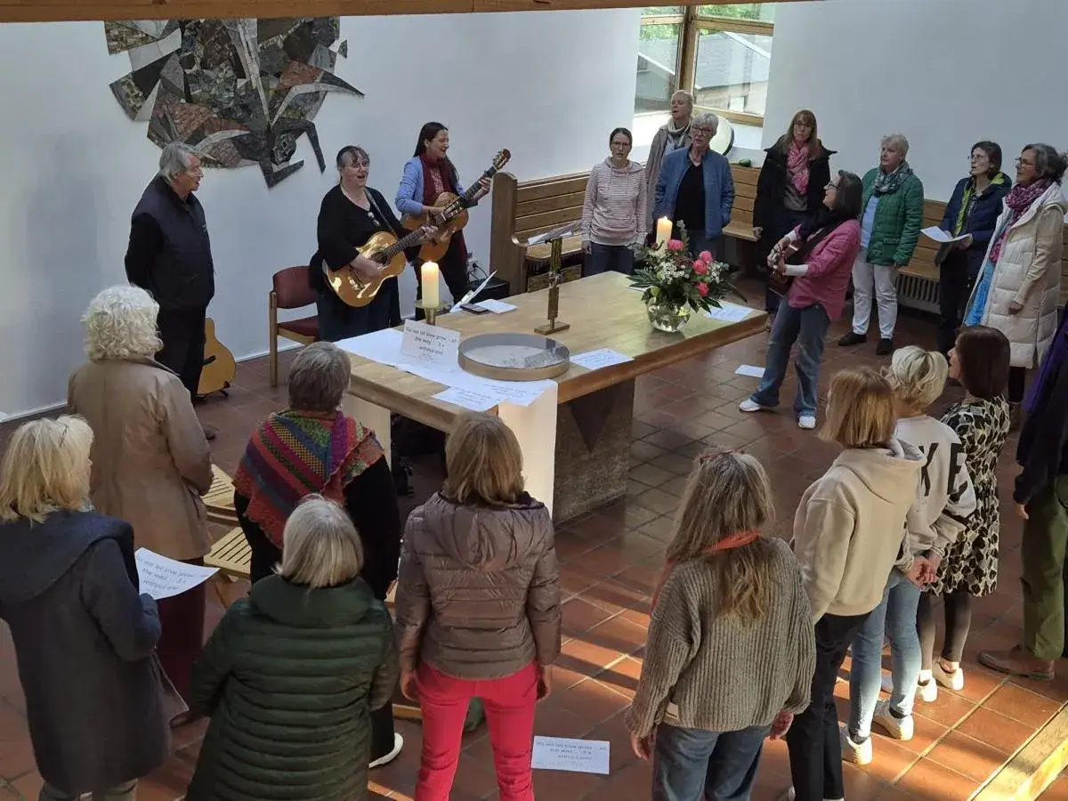 Menschen singen und musizieren in einer Kirche. Eine Frau spielt Gitarre und singt, w&auml;hrend eine andere sie begleitet. Eine Gruppe von Menschen h&ouml;rt zu und singt mit. Ein sch&ouml;ner Moment des gemeinsamen **chant deutsch**.