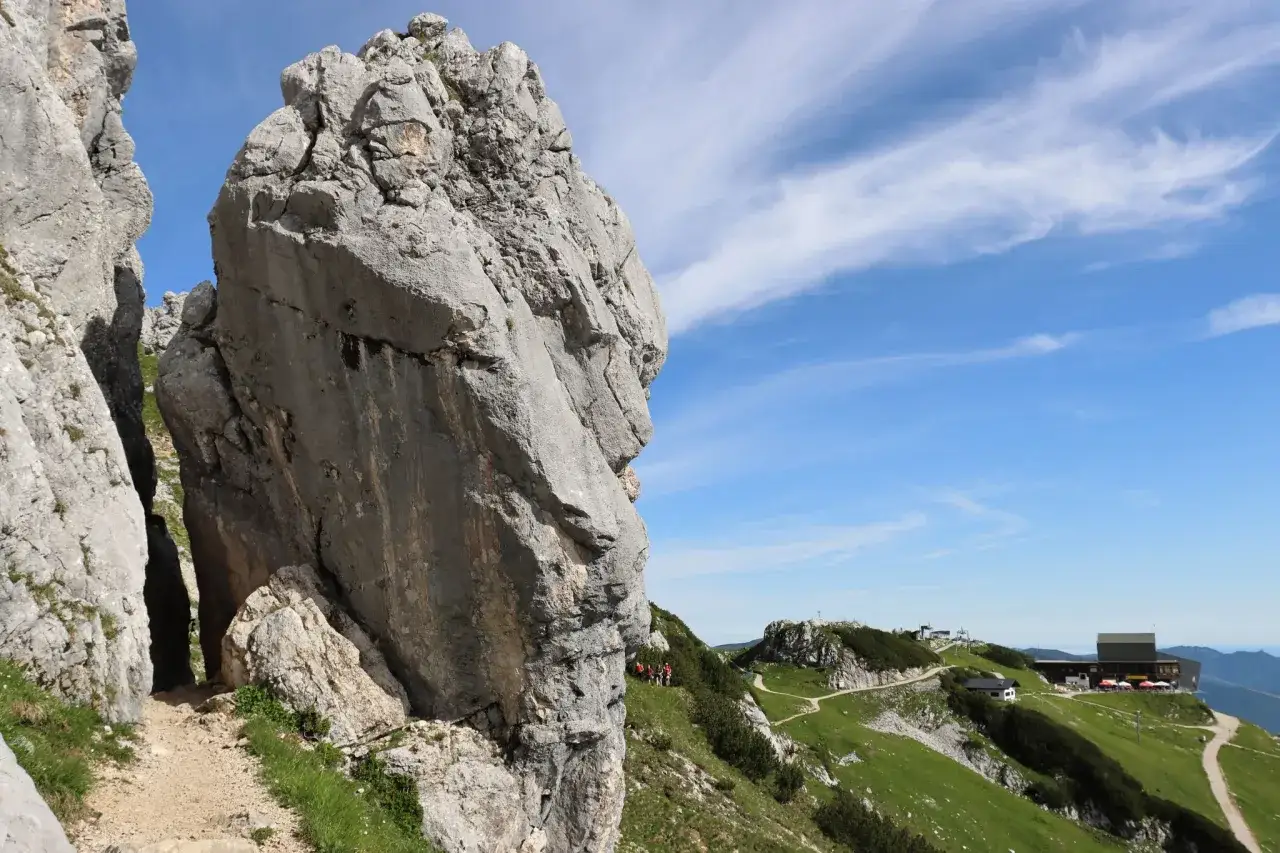 Steiler Felsweg auf der Alpspitz Ferrata. Wanderer genie&szlig;en die Aussicht auf gr&uuml;ne H&auml;nge und ein Berggasthaus.