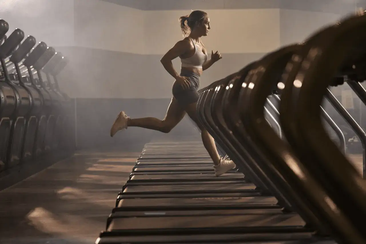 Kobieta w sportowym stroju wykonuje intensywny trening na bieżni, w tle rzędy innych maszyn.