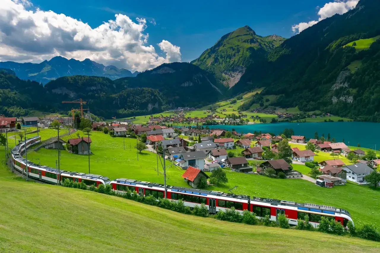 A red and white train winds through a green Swiss village nestled beside a turquoise lake, mountains in the background. This view is perfect for a Switzerland train pass adventure.
