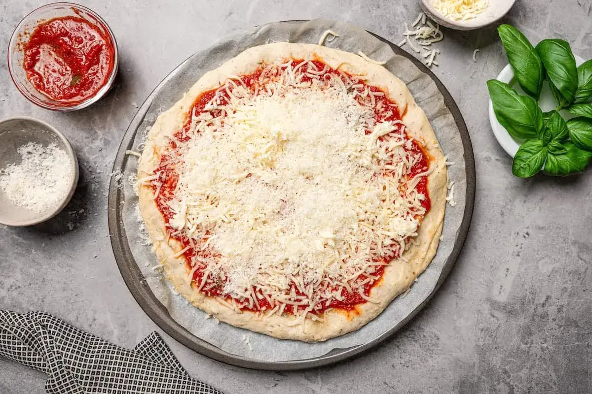 Niegotowa pizza z sosem pomidorowym i obficie posypana startym serem, gotowa na dodatek serka wiejskiego. Obok miseczka z sosem i bazylia.