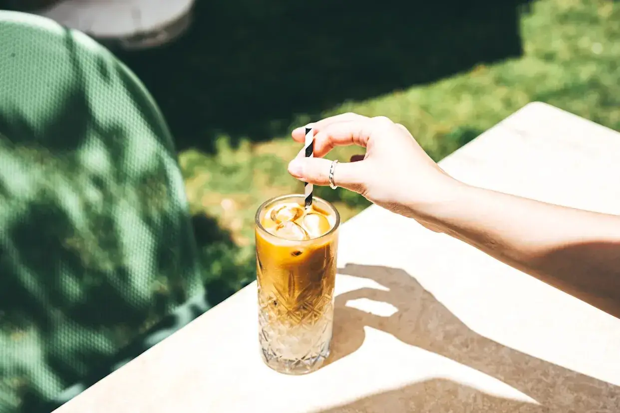 Erfrischender Frappe Espresso mit Eiswürfeln, der von einer Hand mit einem gestreiften Strohhalm gehalten wird.