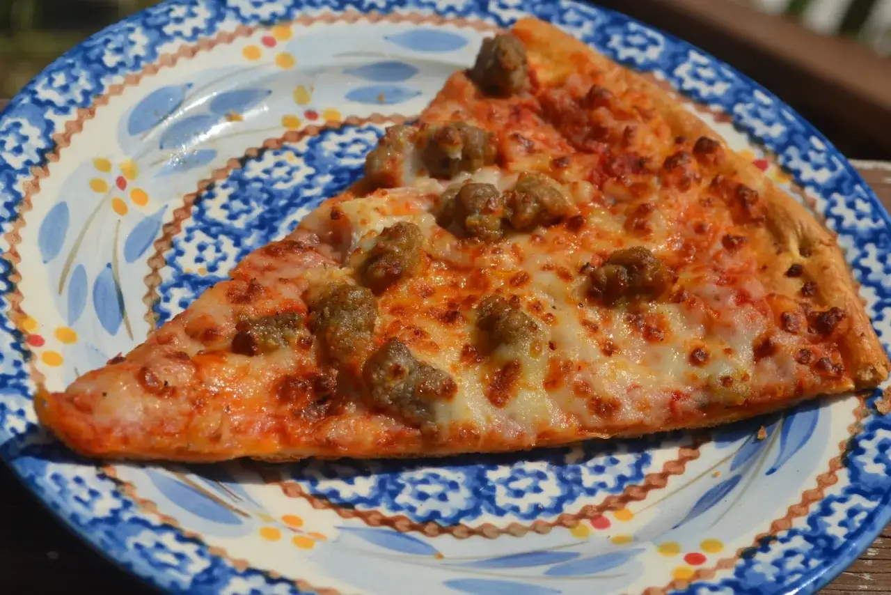 Ein St&uuml;ck Pizza mit Wurstbelag, bereit zum Pizza aufbacken, liegt auf einem blauen Teller mit Muster.