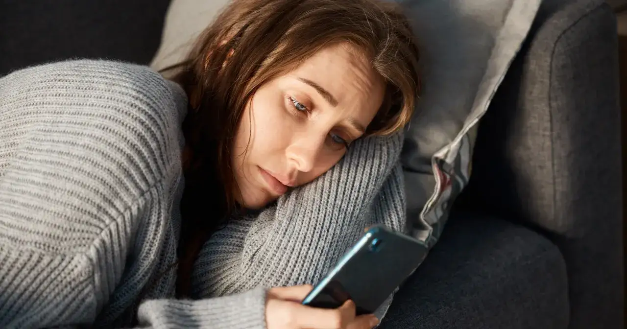 Smutna kobieta w szarym swetrze leży na kanapie, wpatrując się w telefon. Jej oczy wyrażają zmęczenie i pustkę, być może to objawy uzależnienia od ekranu.