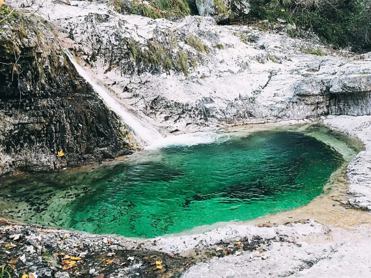 Cascata e pozza smeraldo nella valle del Mis, con rocce bianche e vegetazione.