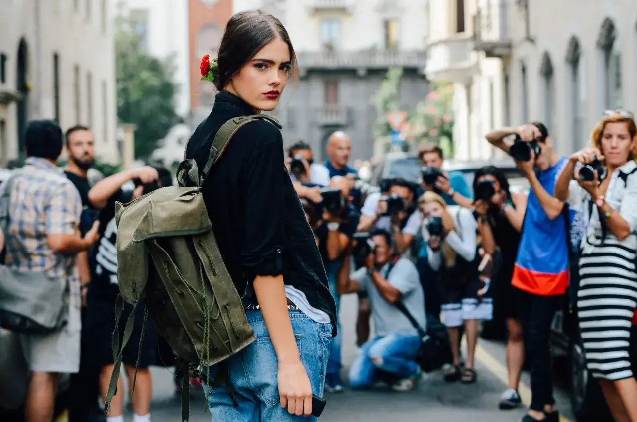 A young woman with a red flower in her hair and a vintage backpack exemplifies the backpacks trend, as paparazzi surround her.